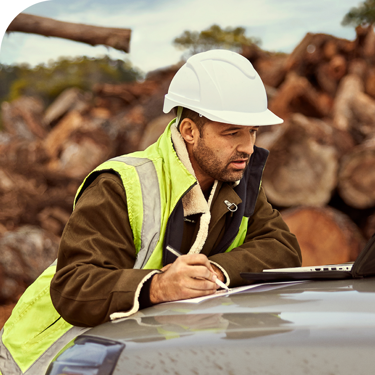 man leaning on car bonnet looking over building plans wearing a hard hat