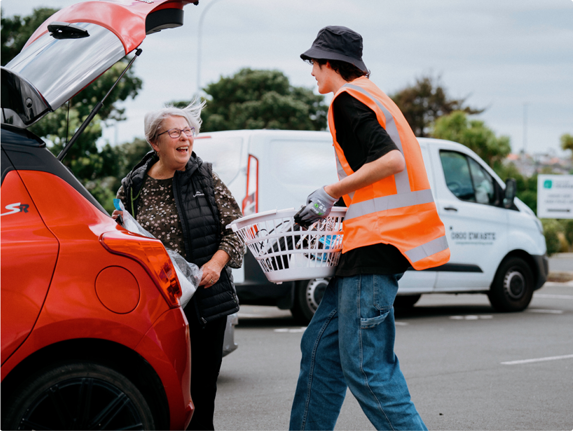 Photo of a young man wearing hi vis carrying a washing barket full with items from the boot of a ladies red car