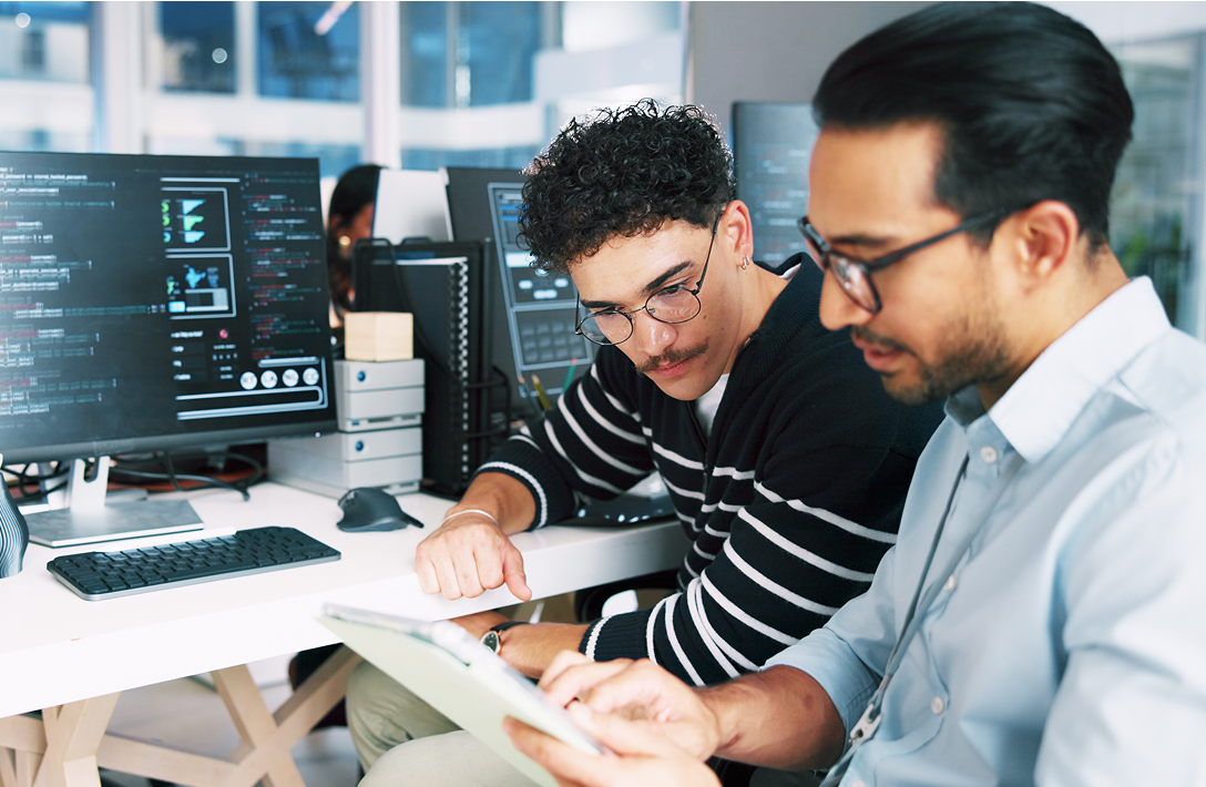 Image of two men sitting at a desk working together with laptops, desktops and a tablet